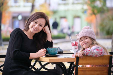 Young pretty woman having good time with her child daughter sitting at street cafe with hot drinks on sunny autumn day. Happiness in family relations conceptの写真素材