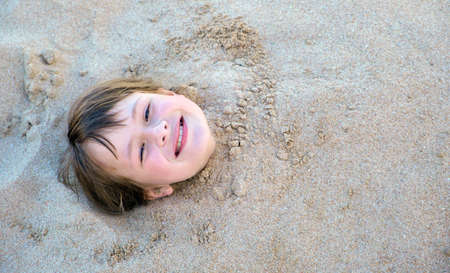 Young smiling child girl lying down covered with white sand on tropic beach on blue sky and ocean...