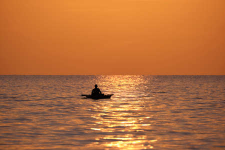 Dark silhouette of lonely fisherman rowing on his boat on sea water at sunsetの写真素材