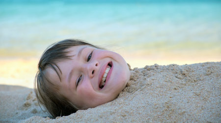 Young smiling child girl lying down covered with white sand on tropic beach on blue sky and ocean water backgroundの写真素材