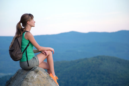 Sportswoman sitting alone taking a break on hillside trail. Female hiker enjoying view of evening nature from rocky cliff on wilderness path. Active lifestyle conceptの写真素材
