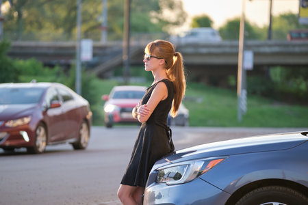 Young woman driver enjoying warm summer day standing beside her car on city street. Travelling and...