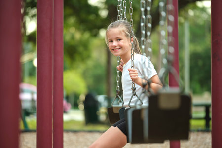 Young pretty happy smiling teenage small girl having fun on playground swings on summer vacations sunny dayの写真素材