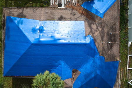 Hurricane Ian damaged house rooftop covered with protective plastic tarp against rain water leaking until replacement of asphalt shingles. Aftermath of natural disasterの写真素材