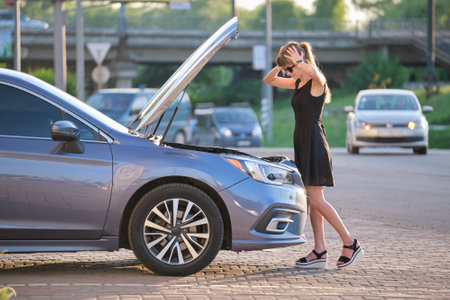Puzzled female driver standing on a city street near her car with popped up hood looking at broken...