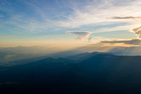 Aerial view of dark mountain hills with bright sunrays of setting sun at sunset. Hazy peaks and...