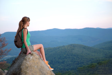 Hiker girl resting on rocky mountain top enjoying summer nature during her travel on wilderness trail. Lonely female traveler traversing high hilltop route. Healthy lifestyle conceptの写真素材