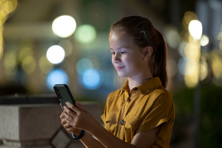 Positive child girl playing online game on her mobile phone with a friend outdoors at nightの写真素材