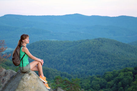 Sportive woman sitting alone taking a break on hillside trail. Female hiker enjoying view of nature from rocky cliff on wilderness path. Active lifestyle conceptの写真素材