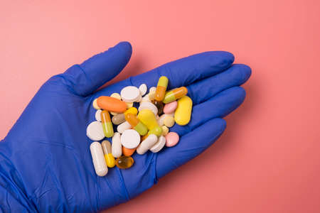 Close up of doctor hand in blue glove and many different pills on pink background.
