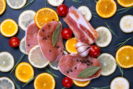 Raw meat stakes, red cherry tomatoes, orange and lemon slices and green salad leaves on dark black background.の写真素材