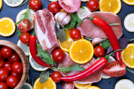Raw meat stakes, red cherry tomatoes, orange and lemon slices and green salad leaves on dark black background.の写真素材