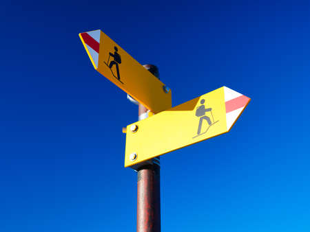 Directional marker for a mountain hike. Yellow bright sign with an arrow against the blue sky. Navigation in a hiking trip. High resolution photo.の写真素材