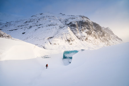 A man in front of the entrance to an ice cave in Iceland. A walk on the glacier. High mountains and clouds at dawn. Travelling through Iceland in a Vatnajokull National Park.の写真素材