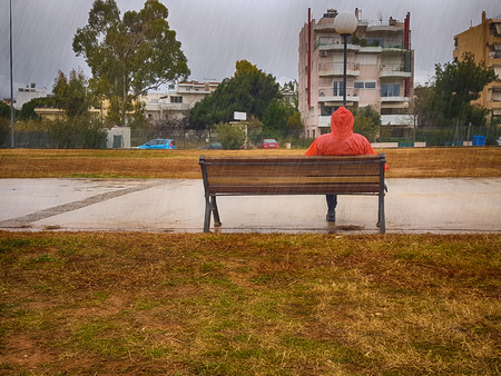 Stranger with a red coat sitting on a bench at a park on a rainy day against a dramatic sky.の写真素材
