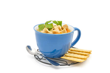 Hearty chicken noodle soup served with crackers in a blue ceramic cup on a white background.の写真素材