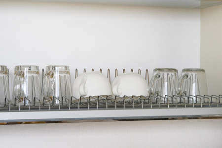 Washed white glass and ceramic cups on metal rack inside kitchen cupboard. Modern kitchen with tableware dryingの写真素材