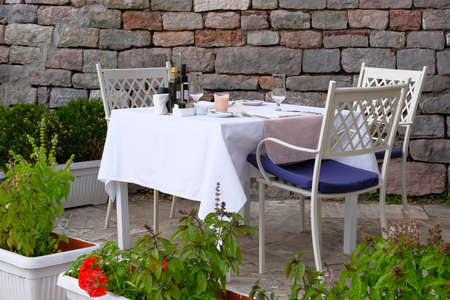 Table with white tablecloth, cutlery and wine glasses on outdoor summer veranda of restaurant. Wall of old textured stone in background.の写真素材