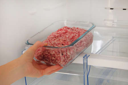 Minced raw meat in glass container is on shelf in open fridge. Female hand pulls out from fridge an ingredient for cooking meat dishes.の写真素材