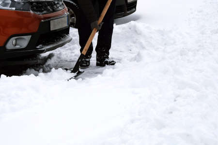 Man brushing and shoveling snow from orange car in winter after snowfall. Shovel in hand. Winter problems of car drivers.の写真素材