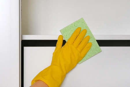 Housewife cleans the house, wipes dust with a cleaning rag. Hand in yellow protective glove is...