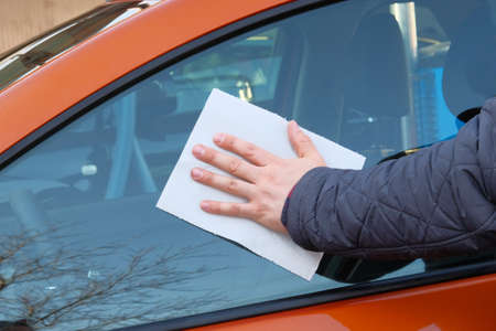 Man after washing wipes glass window with a white paper napkin. Male hand and car body close up. Orange car at car wash.の写真素材