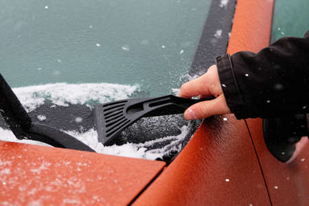 Scraper in mans hand. Man clears snow from icy windshield of car. Cleaning frozen window of orange automobile. Snowy.の写真素材