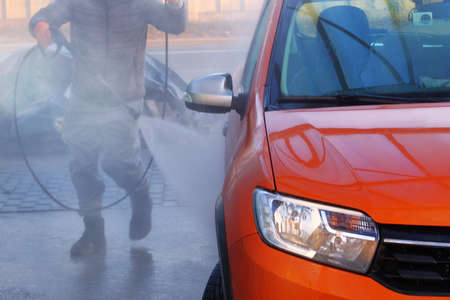 Man washes his orange car at car wash in outdoors. Cleaning with with a water jet at self-service car wash. Soapy water runs down.の写真素材