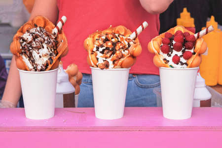 Bubble waffles with raspberries, chocolate and marshmallow in white paper cups are sold on food festival event. Street sweet food in food stall.の写真素材