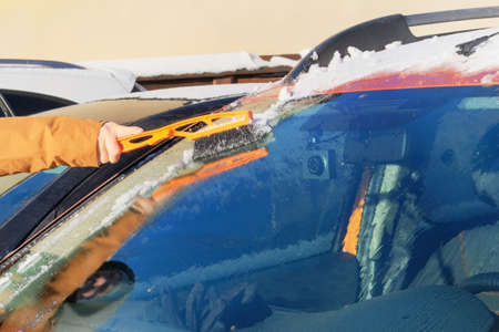 Man clears orange car from snow. Windshield of car. Brush in mans hand. Winter, snow, vehicle.