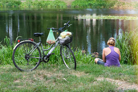 Young girl is resting in a park near a lake after riding her bicycle to the grocery store. Shopping by bike.の写真素材