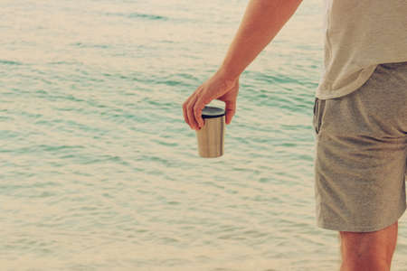 Tourist food for outdoor activities. Metal thermo cup in hand on coastline. Enjoy a tasty drinks...