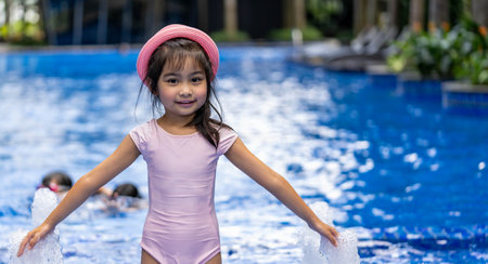 Portrait of pretty asian child smilling and posing on swimming pool background wearing pink swim suit and pink summer hatの写真素材