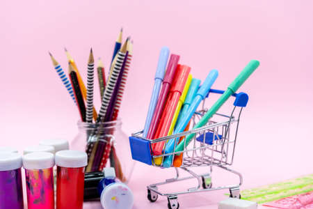 Bright stationery objects in mini supermarket cart on pink background. Back to school concept.
