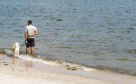 Man while walking his dog on a beach side with white sand and wavesの写真素材