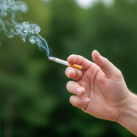 A close-up shot of a hand holding a lit marijuana joint, with smoke curling upwards against a blurred green background.の素材