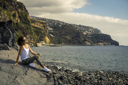 Young and attractive girl in white shirt and sneakers is sitting on the rocky beach on Madeiras island. Beautiful landscape on background. Sunny day.の写真素材