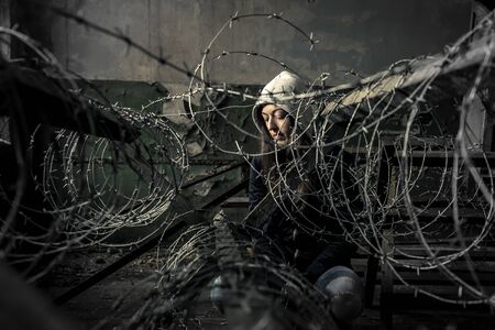 Young girl is sitting behind barbwire in old, ruined factory.