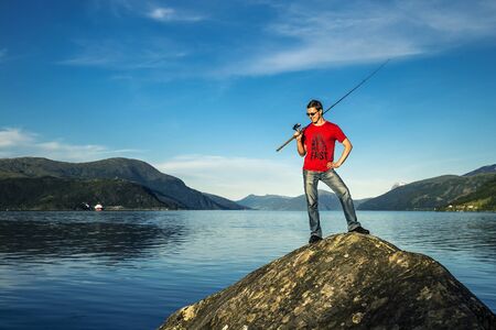 Young man is fishing in Norway. He is standing on the stone and smiling.の写真素材