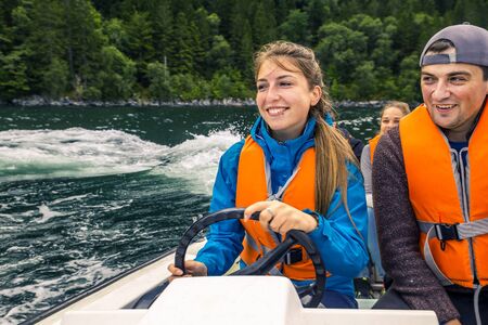 Portrait of young and attractive woman close up in blue jacket and orange safe jacket sitting in a boat with raised up hand and smiling, Norway.の写真素材