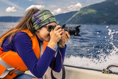 Portrait of young and beautiful woman sitting in a boat and looking at her camera. Water splashes on background. Beautiful sky on background, Norwayの写真素材