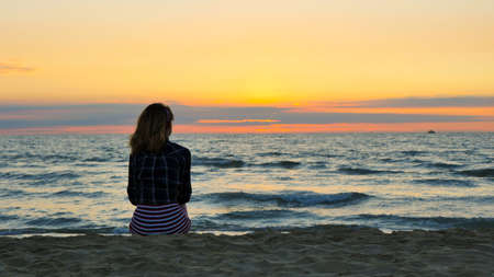 Blonde girl relaxing on the beach and watching the sunset. Moment of meditation. Sea, waves and orange sky in the backgroundの写真素材