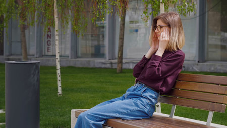 Woman suffering bad headache touching her temples outside sitting on a bench outdoor. Portrait of sad young Caucasian. Migraine. Unhappy face.の写真素材
