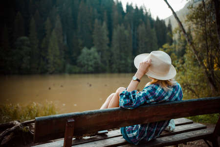 Young woman in a denim shirt and hat, sitting on a bench, admires the lake landscape, the pine forest and the relief of the mountains in the backgroundの写真素材
