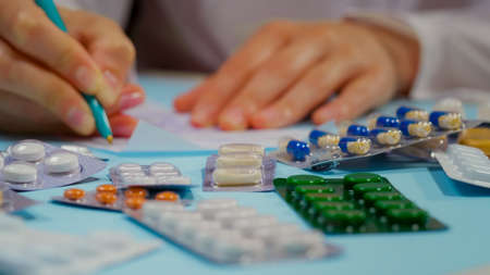 Doctor hand writing a prescription. Nearby Pile of pills in blister packs on blue background. Close Up.の写真素材