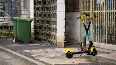 Two electric scooters on the sidewalk behind a block next to a trash can