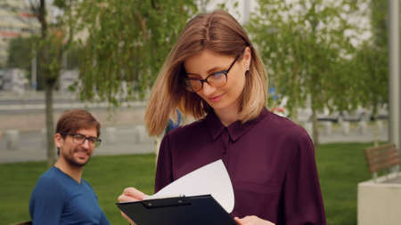 Confident Businesswoman walking out of Business building. Young blonde Woman greets friend sitting on a bench while the Walk. Success. Happy woman expression.の写真素材
