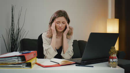 Tired woman, overwhelmed by laptop work, has severe headache. She takes a break while massaging her temples, is tired and feels unwell but tries to concentrate. Overworked woman with migraine.の写真素材