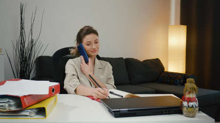 Young busy business woman working on project paperwork talking on the phone with partner. Female manager making business notes on notebook, working alone in office.の写真素材