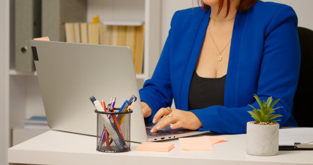 Office worker typing report on laptop at desk in workplace. Woman busy with work.の写真素材